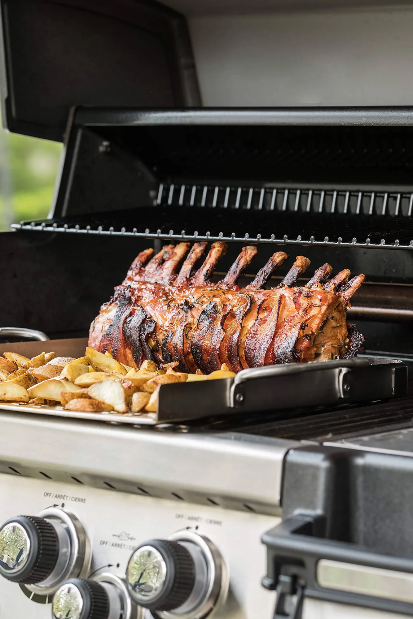Bacon-wrapped rack of pork with roasted potatoes on a gas grill, photo by Alex Slyadnev