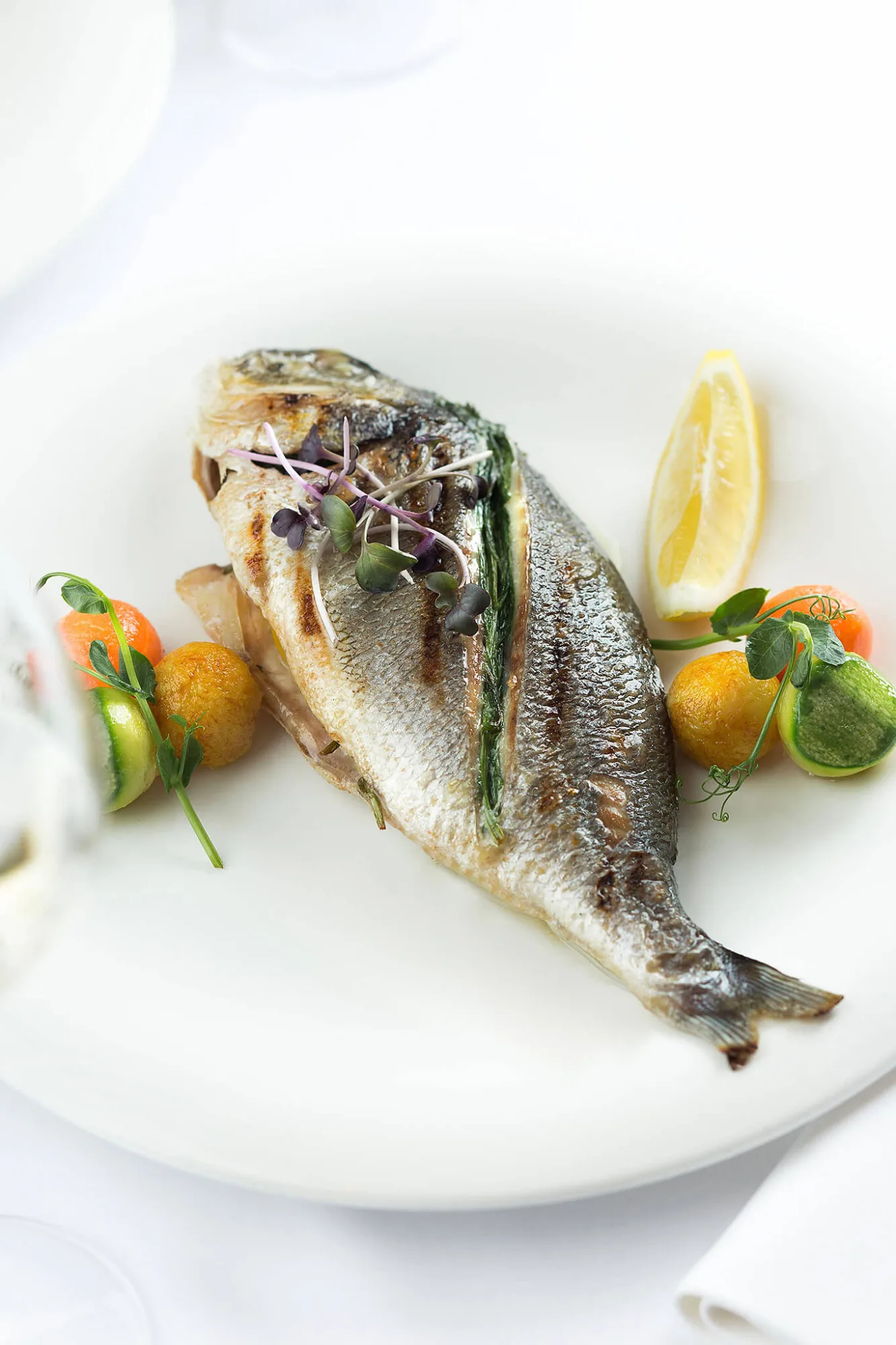 Grilled whole sea bream with lemon and microgreens on a white plate, photo by Alex Slyadnev