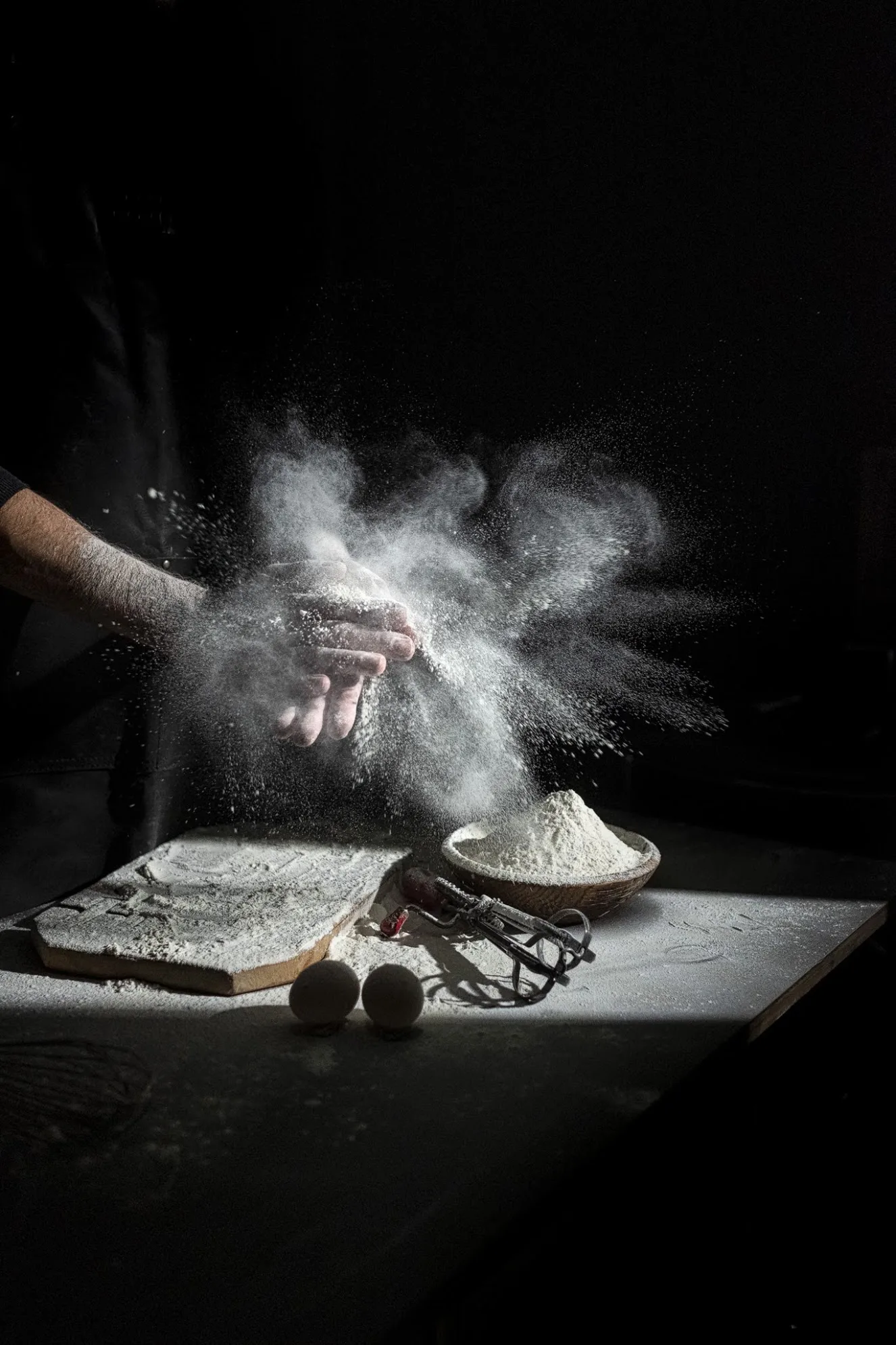 Baker's hands clapping flour over a board with eggs, dark background, photo by Alex Slyadnev